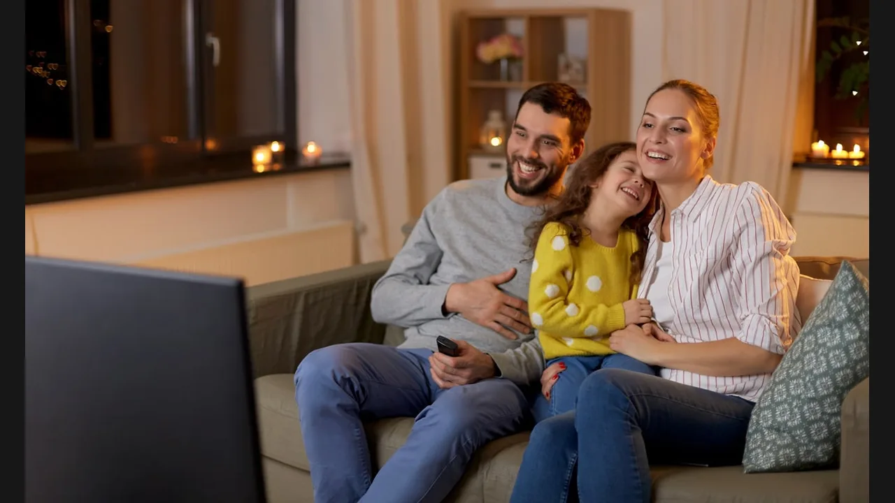 A family on a sofa in a warm living room watching television, the father holding a remote