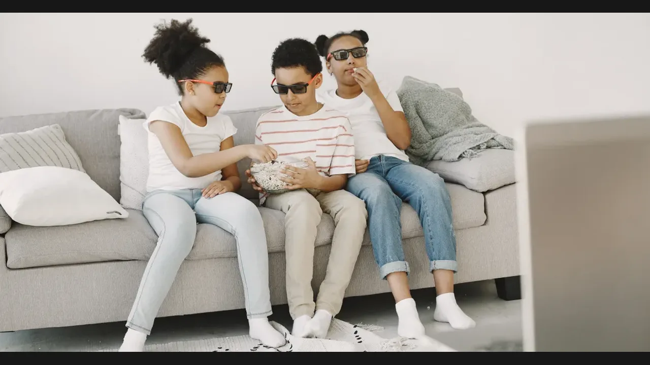 Three children wearing 3D glasses sharing popcorn on a sofa while watching a film