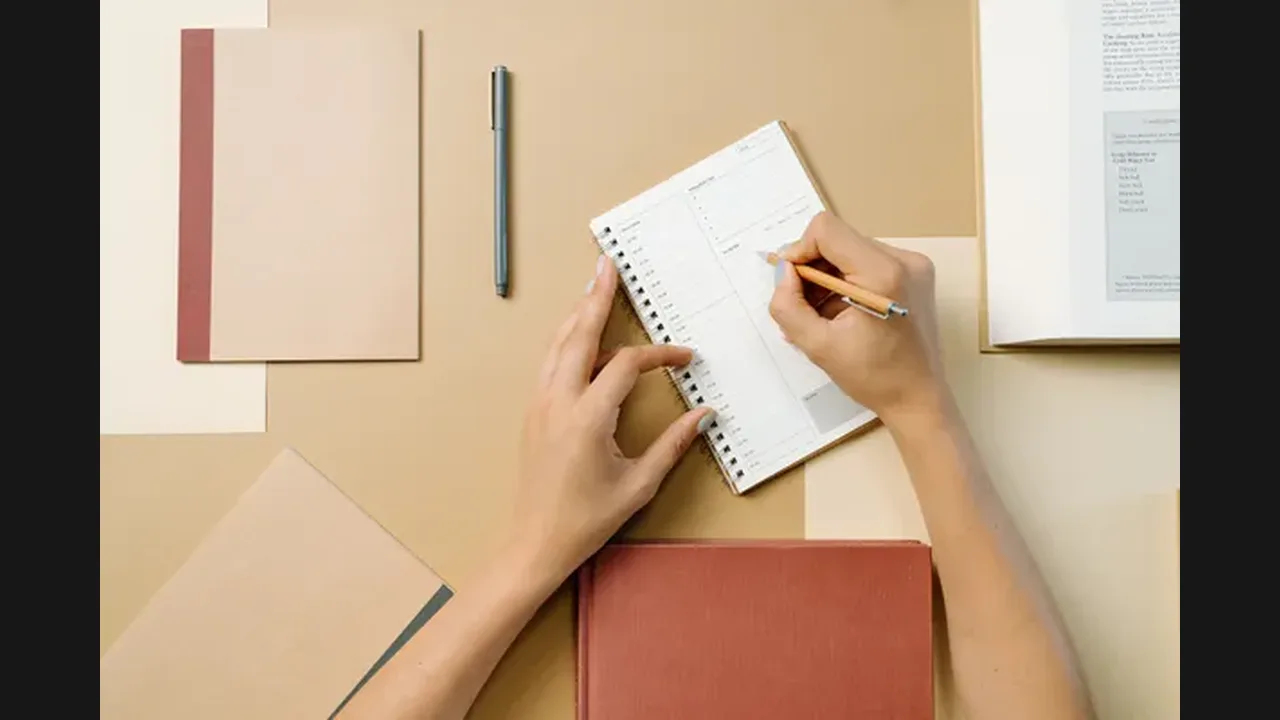 Hands writing in a notebook planner on a desk with books and a pen